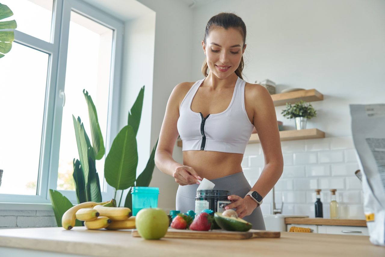 happy young woman in sports clothing preparing protein cocktail at home